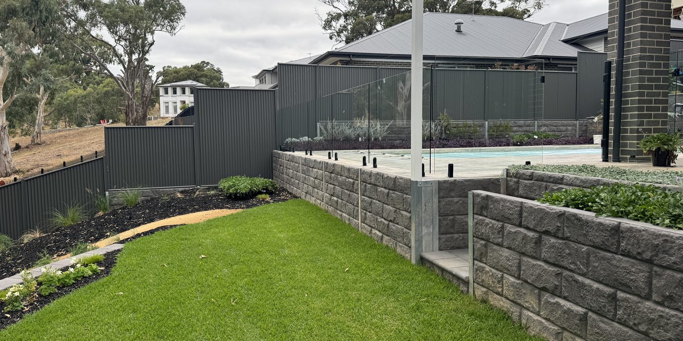 Two tiers of grey stone-textured concrete sleeper retaining walls with galvanised steel posts, a glass pool fence on top and new lawn below, in Craigburn Farm