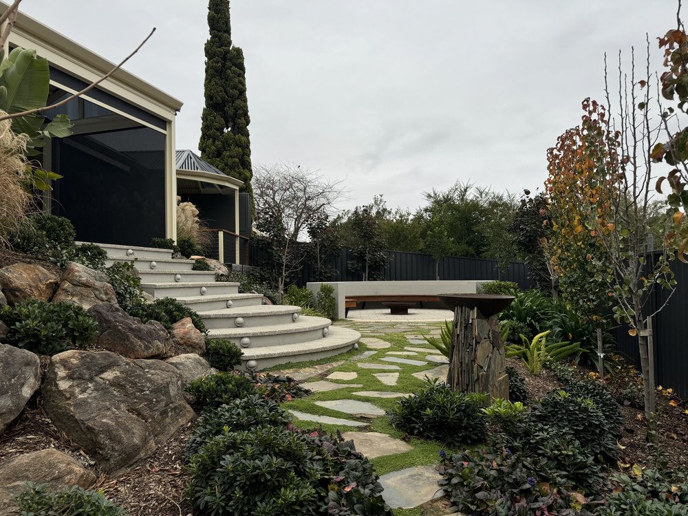 Sloping back garden in Aberfoyle Park with curved steps, boulders, stepping stones set in lawn and a rendered seat wall around the firepit