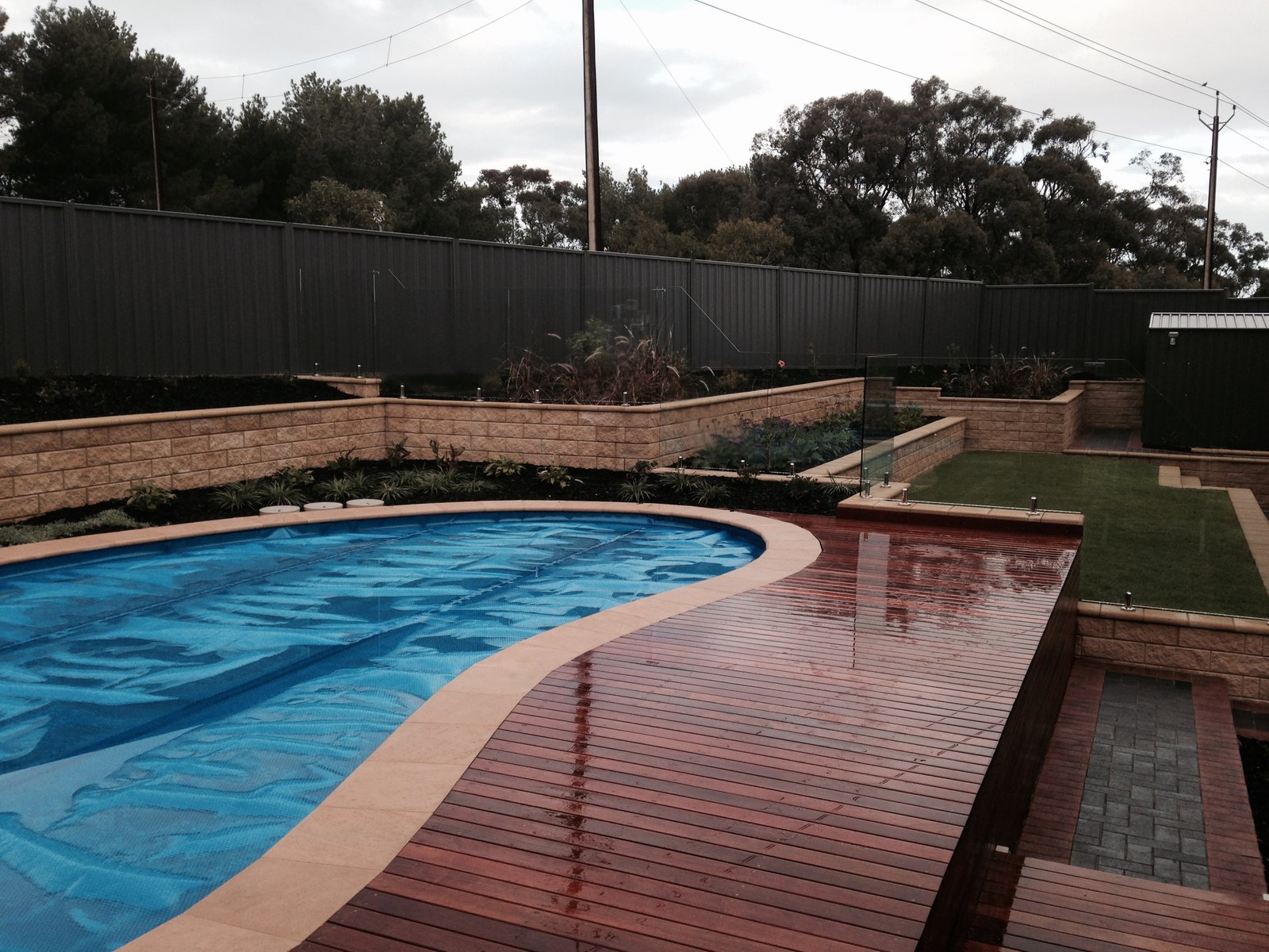 Sandstone-block retaining walls terracing a sloping backyard around an in-ground pool with a hardwood deck and glass pool fencing