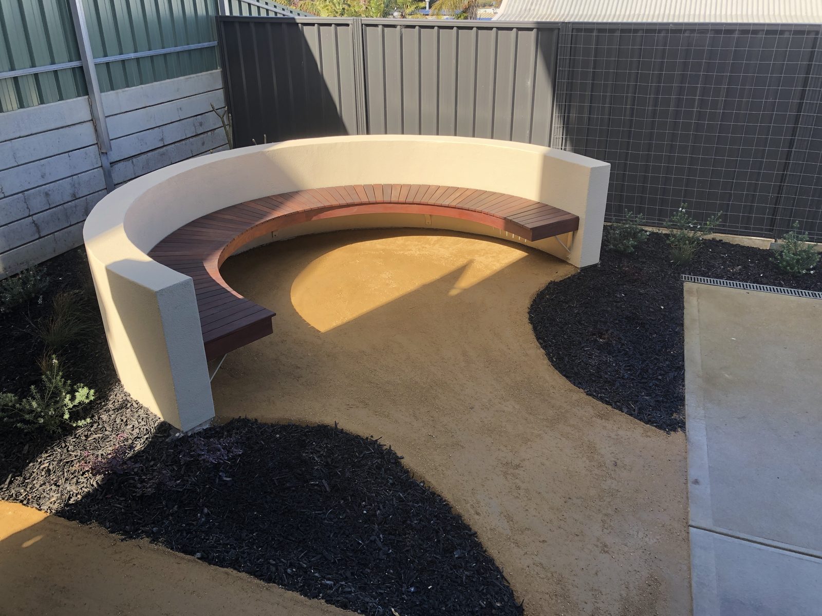 Curved rendered seat wall with hardwood bench seating, surrounded by newly planted native beds under dark mulch in Port Noarlunga