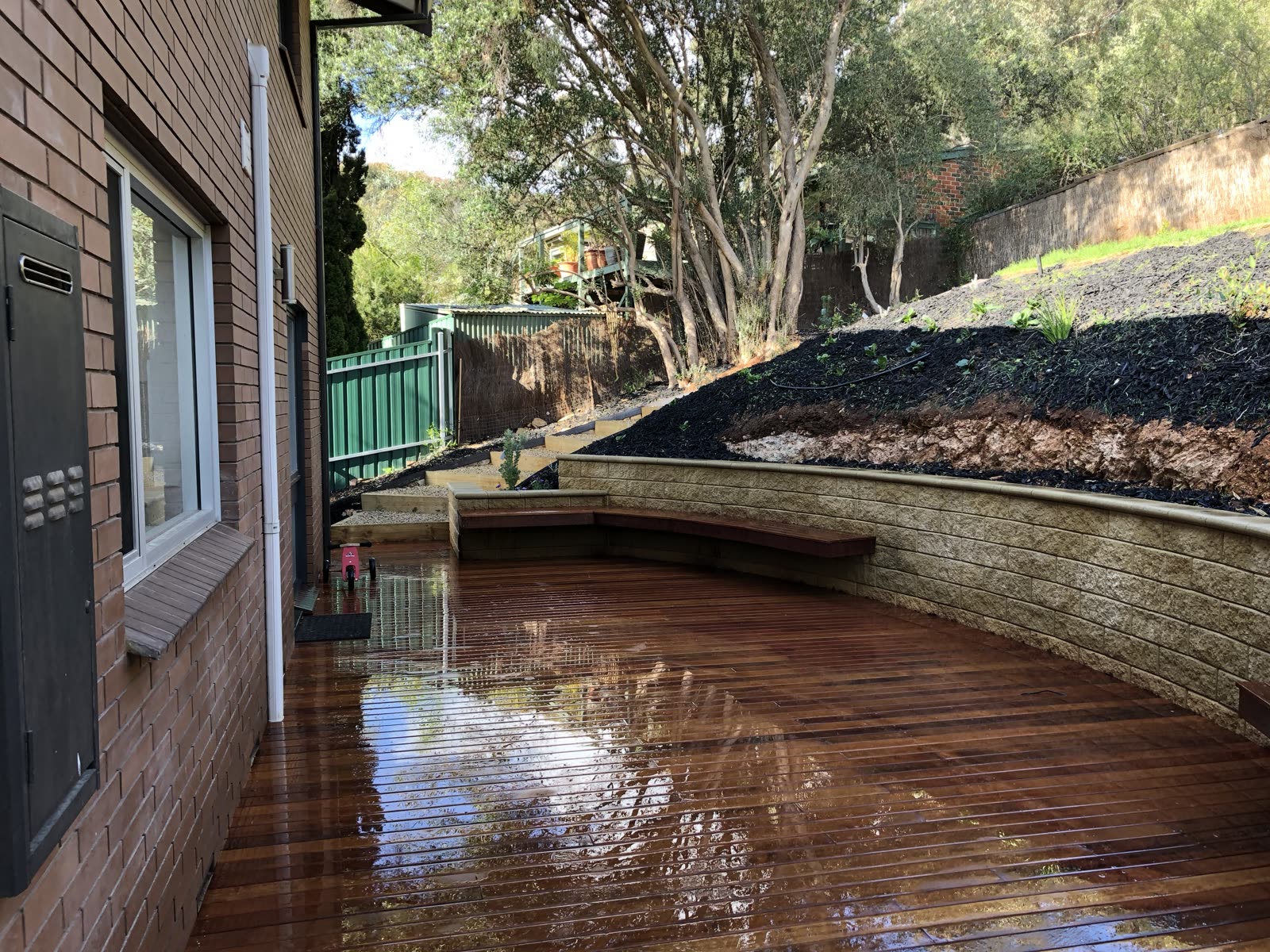 Curved sandstone-block retaining wall behind a hardwood timber deck with a built-in bench, cut into a sloping backyard