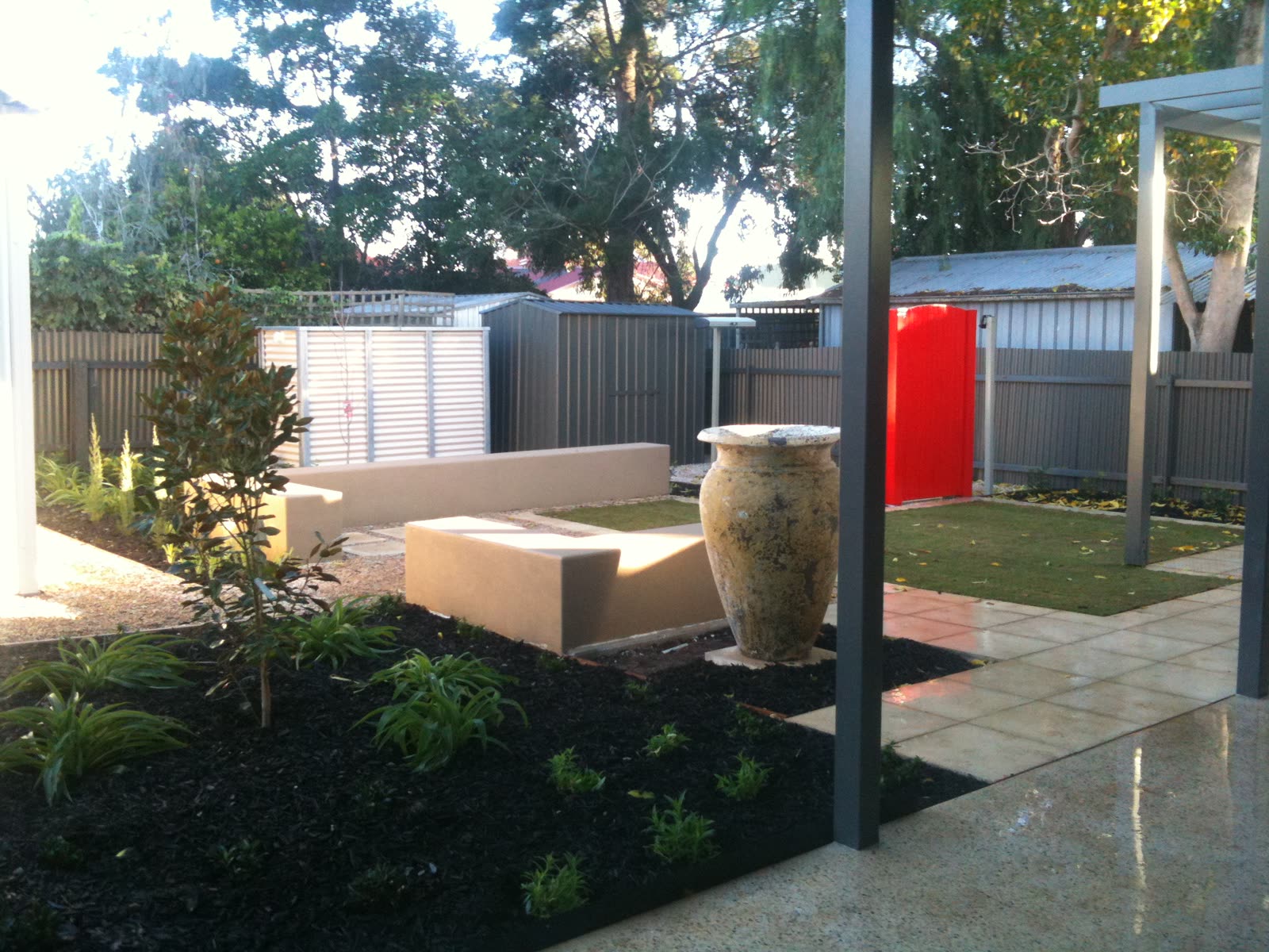 Backyard entertaining area in Westbourne Park with rendered concrete bench seating, a large glazed urn and lawn framed by a pergola